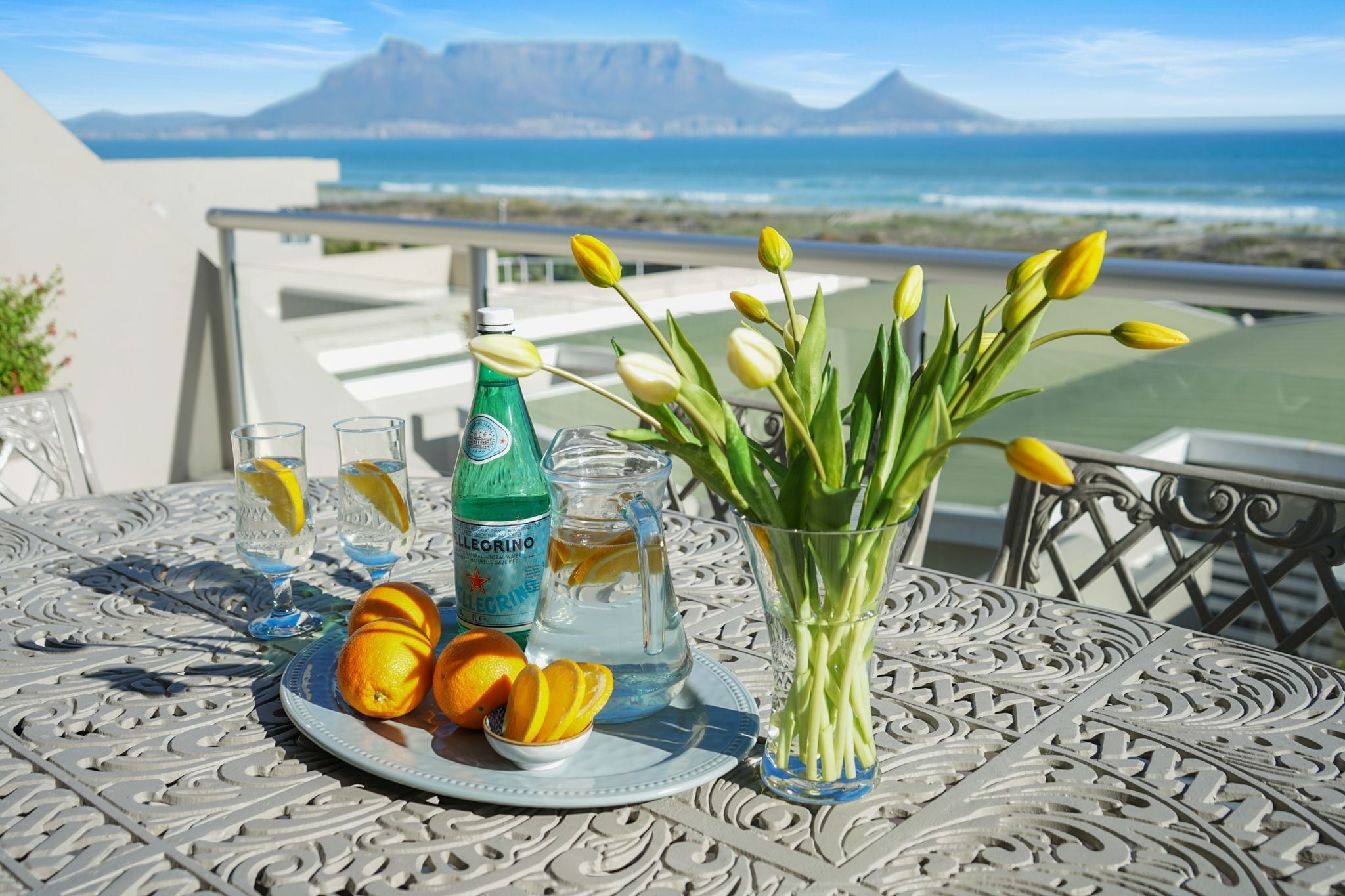 A table with a plate of food, drinks, and a vase of flowers on a balcony with a view of the ocean.