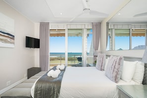 A hotel room with a large bed, a flat-screen TV, a ceiling fan, and a balcony with a view of the ocean. - Dolphin Beach H106 (Cape Town)