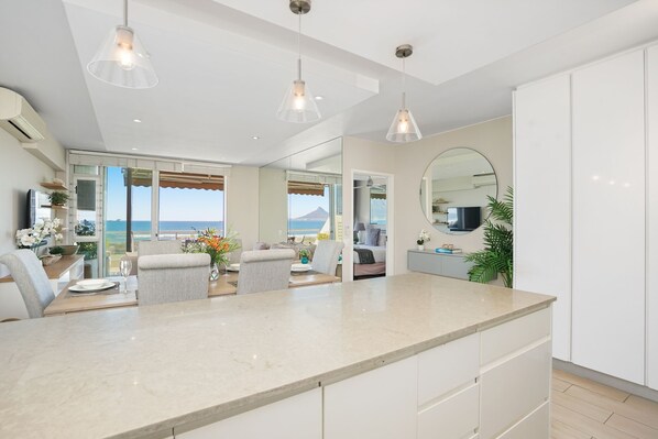 A modern kitchen with a large island, pendant lighting, and a view of the sea. - Dolphin Beach H106 (Cape Town)