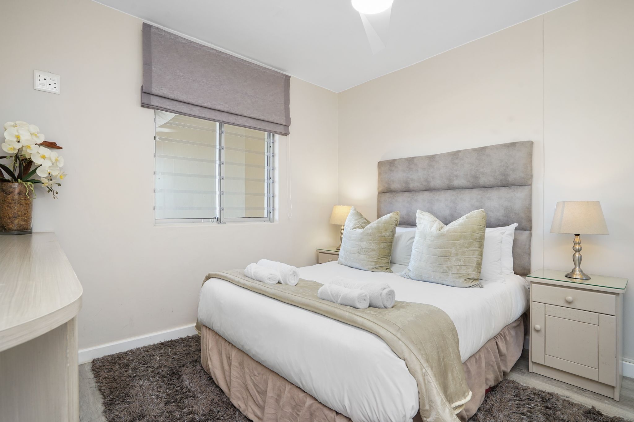 A bedroom with a bed, two bedside lamps, a nightstand, a window with blinds, and a vase with flowers.