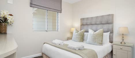 A bedroom with a bed, two bedside lamps, a nightstand, a window with blinds, and a vase with flowers.