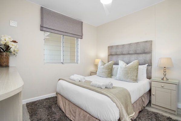 A bedroom with a bed, two bedside lamps, a nightstand, a window with blinds, and a vase with flowers. - Dolphin Beach H106 (Cape Town)