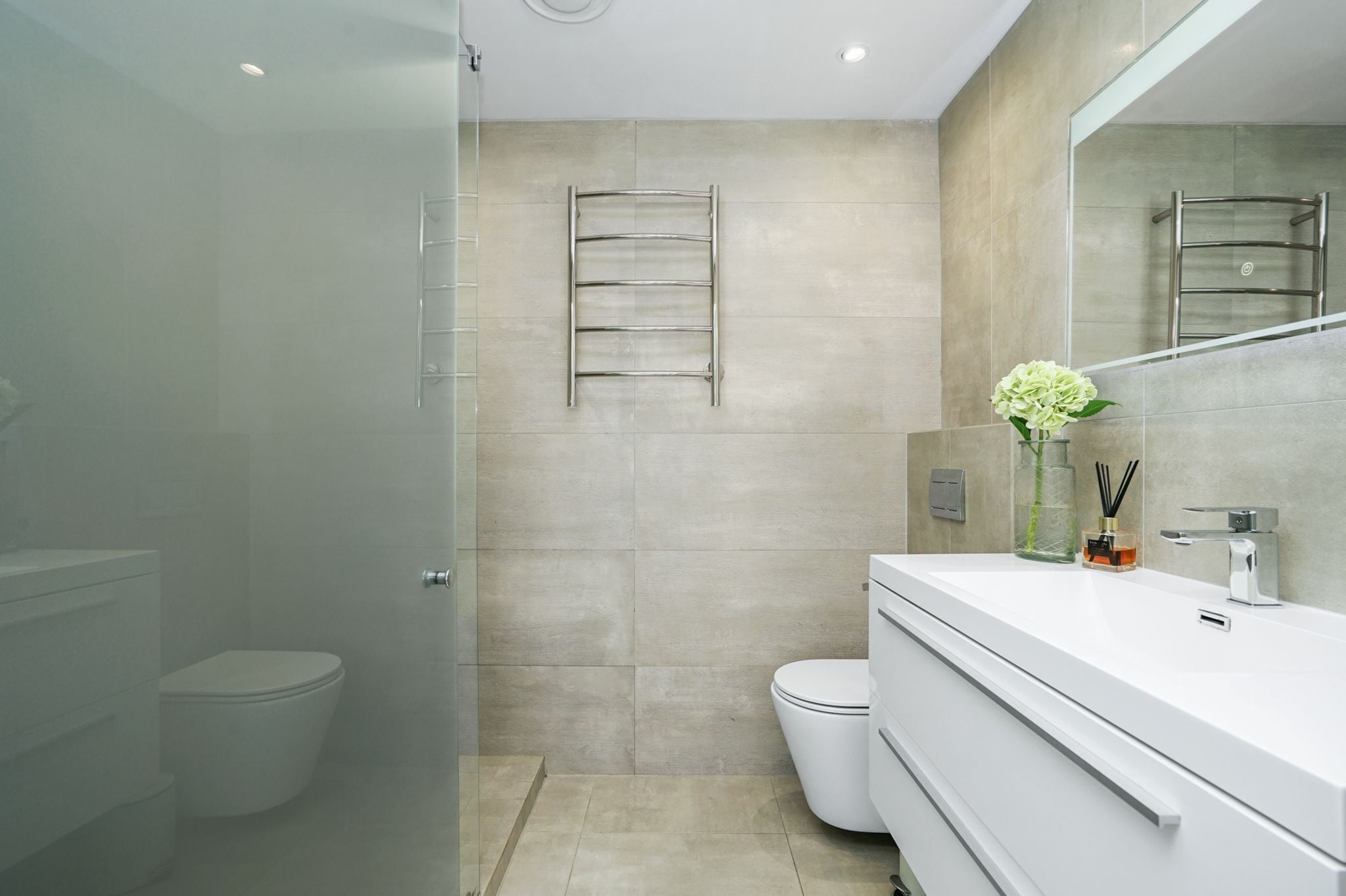 Un baño moderno con cabina de ducha de vidrio, inodoro blanco, tocador blanco con espejo y un perchero en la pared.
