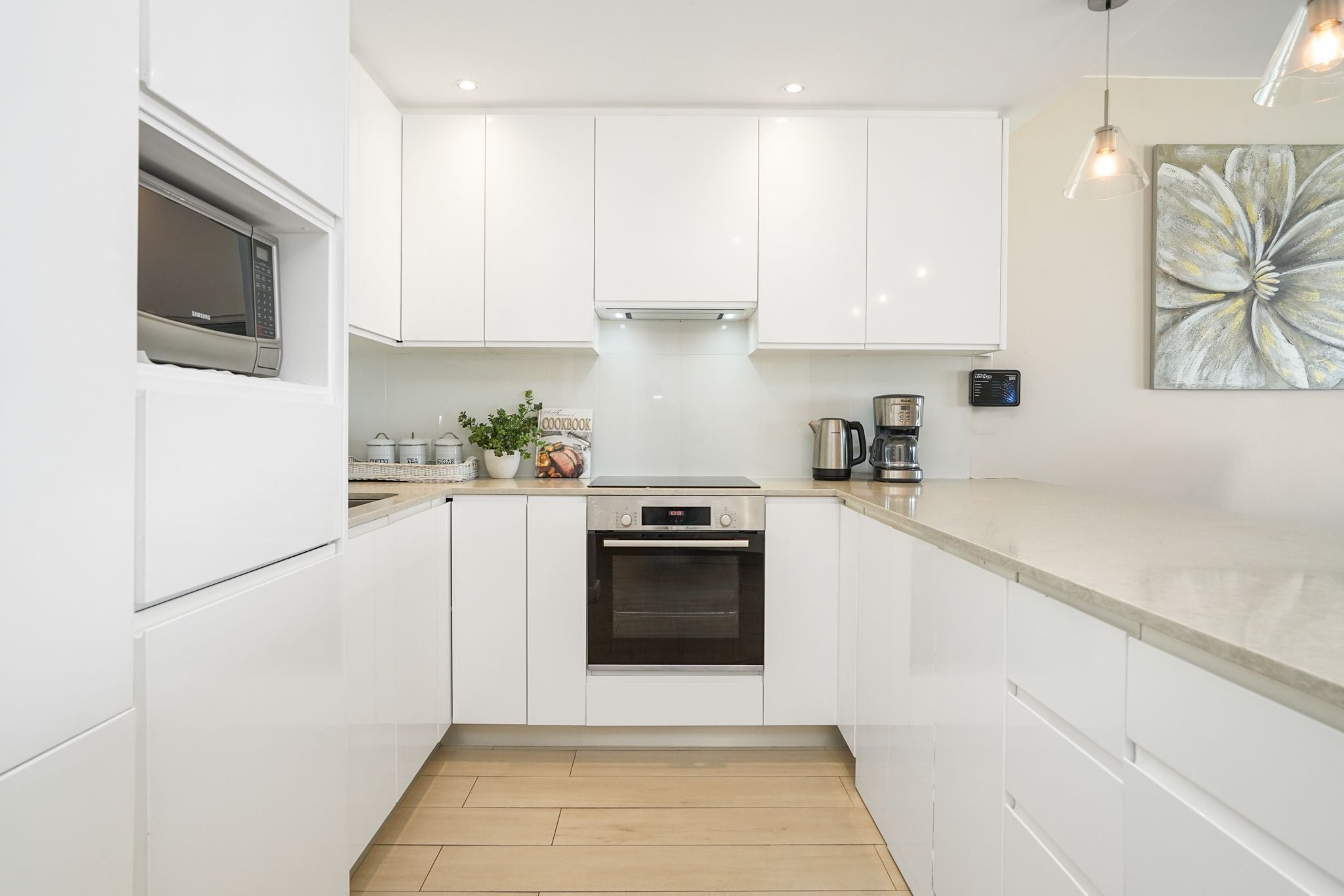A modern kitchen with white cabinets, a built-in oven, a microwave, a coffee maker, and a decorative flower painting on the wall.