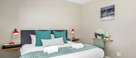 Un lit bien fait, avec une literie blanche et turquoise, deux lampes de chevet, une table de chevet et une étagère murale agrémentée d’un miroir et d’une petite plante.