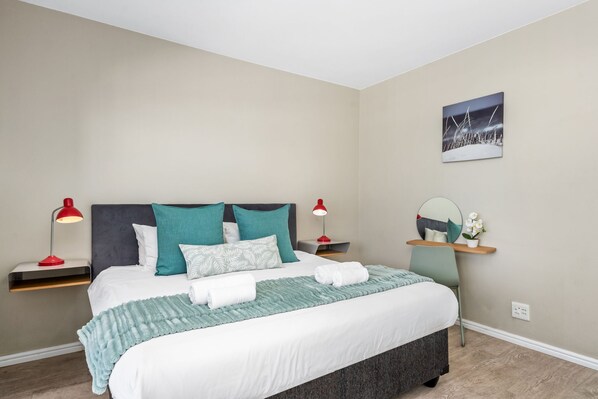 A neatly made bed with white and teal bedding, two bedside lamps, a nightstand, and a wall-mounted shelf with a mirror and a small plant.