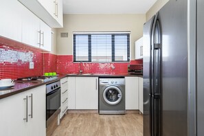 A compact kitchen with white cabinetry, a stainless steel refrigerator, a washing machine, and a window with blinds.