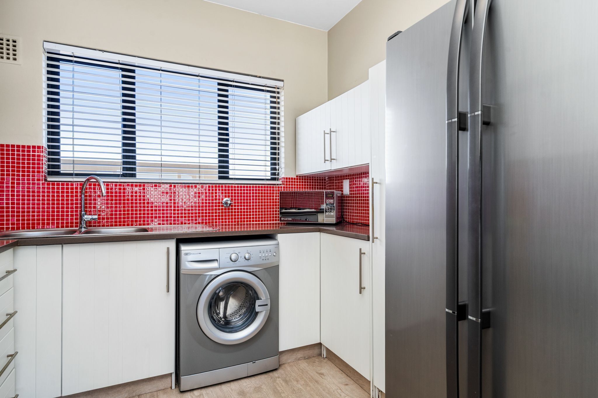 A compact kitchen with a washing machine, a stainless steel refrigerator, and a window with blinds.
