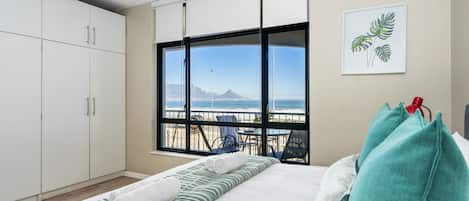 A bedroom with a bed, a window with a view of the beach, a balcony with chairs, and a white wardrobe.