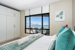 A bedroom with a bed, a window with a view of the beach, a balcony with chairs, and a white wardrobe.