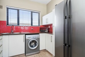 Une cuisine compacte équipée d’un lave-linge, d’un réfrigérateur en acier inoxydable et d’une fenêtre avec des stores.