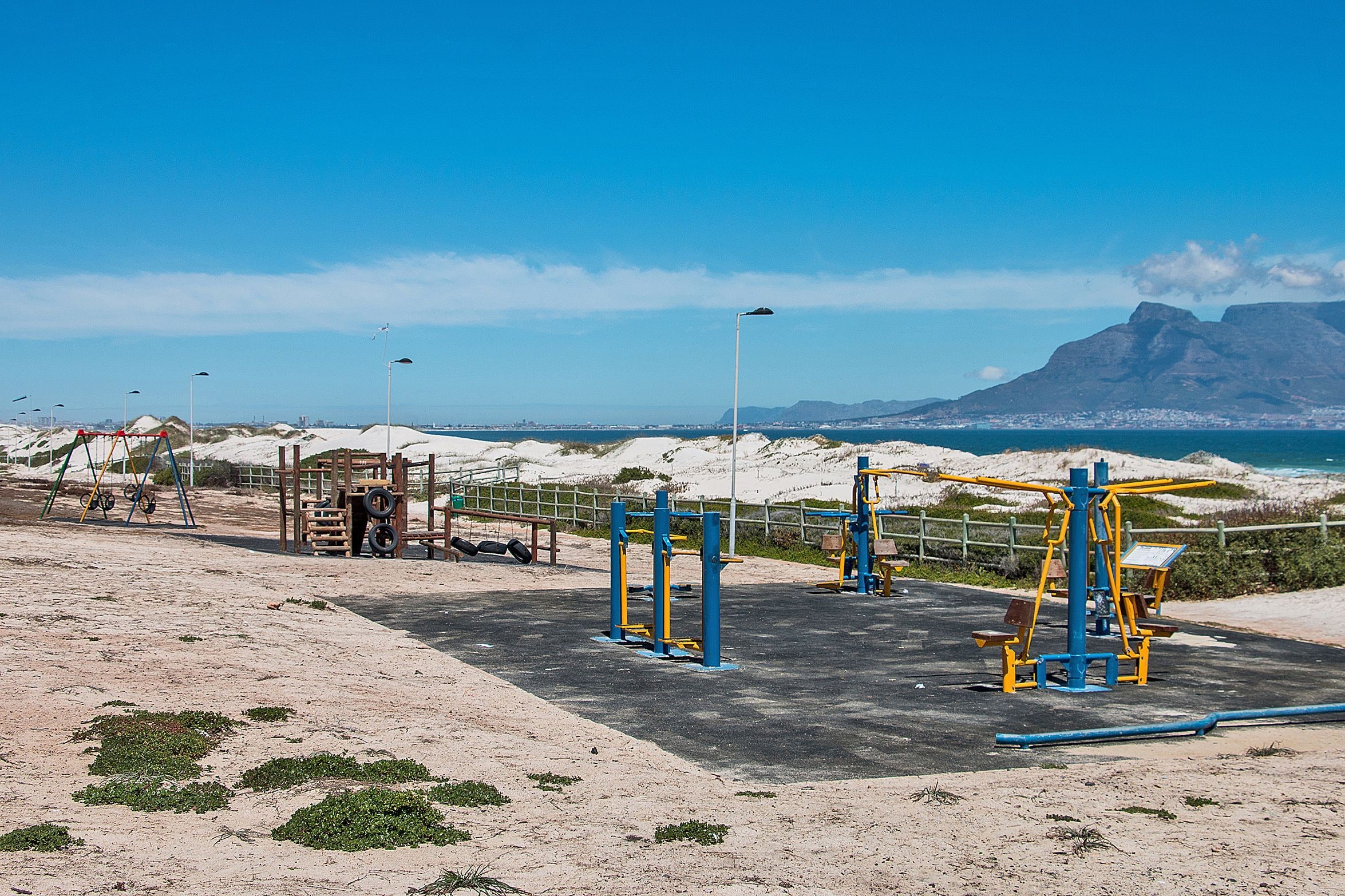 An outdoor gym with exercise equipment, a sandy area, and a view of a mountain and beach in the background.
