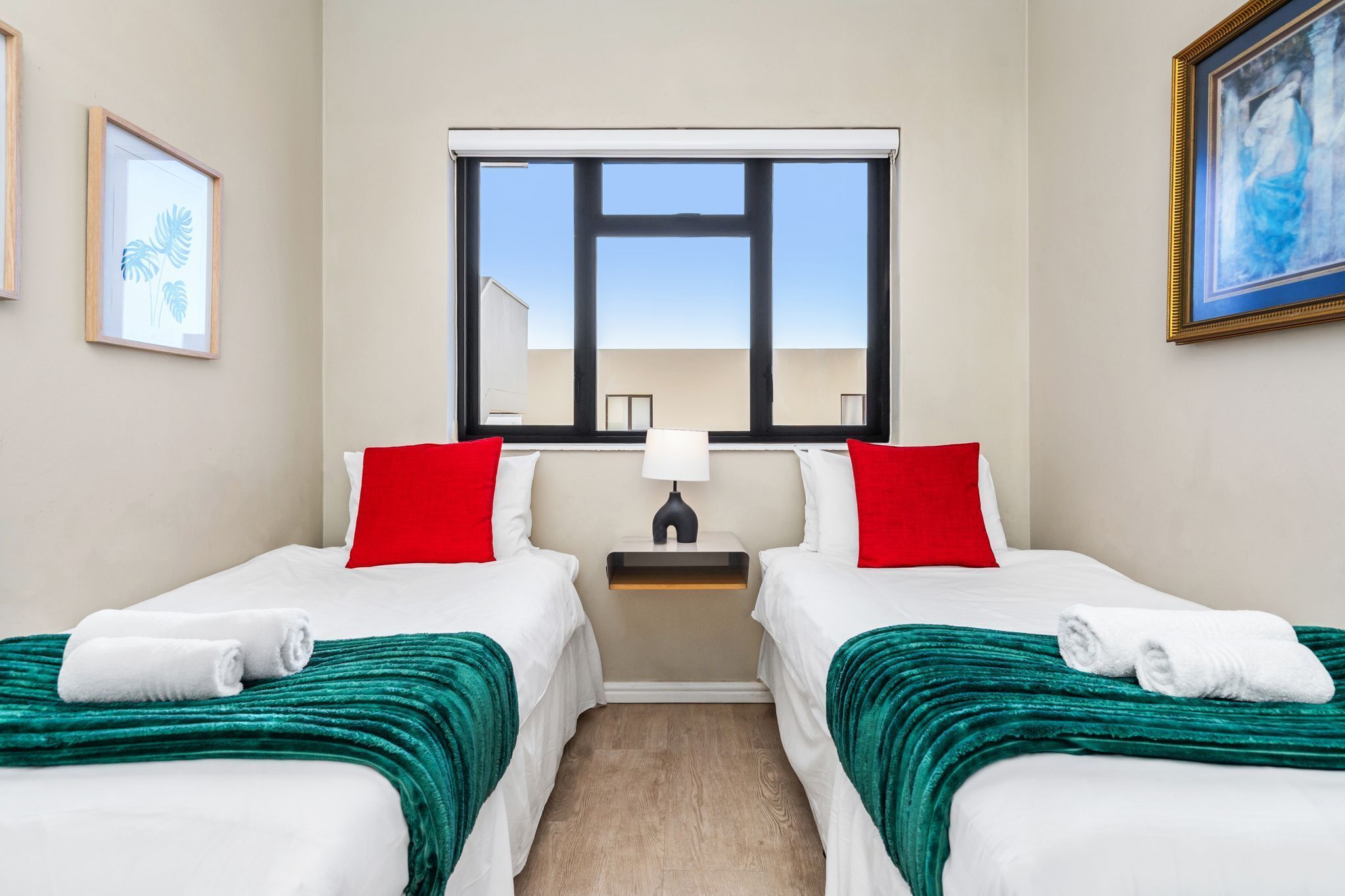 A hotel room with two beds, a window with a view, a nightstand with a lamp, and artwork on the walls.