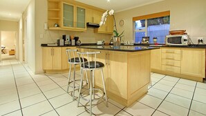 A modern kitchen with wooden cabinets, a central island with bar stools, a microwave, and a window with blinds. - Durham Close 31 (Cape Town)