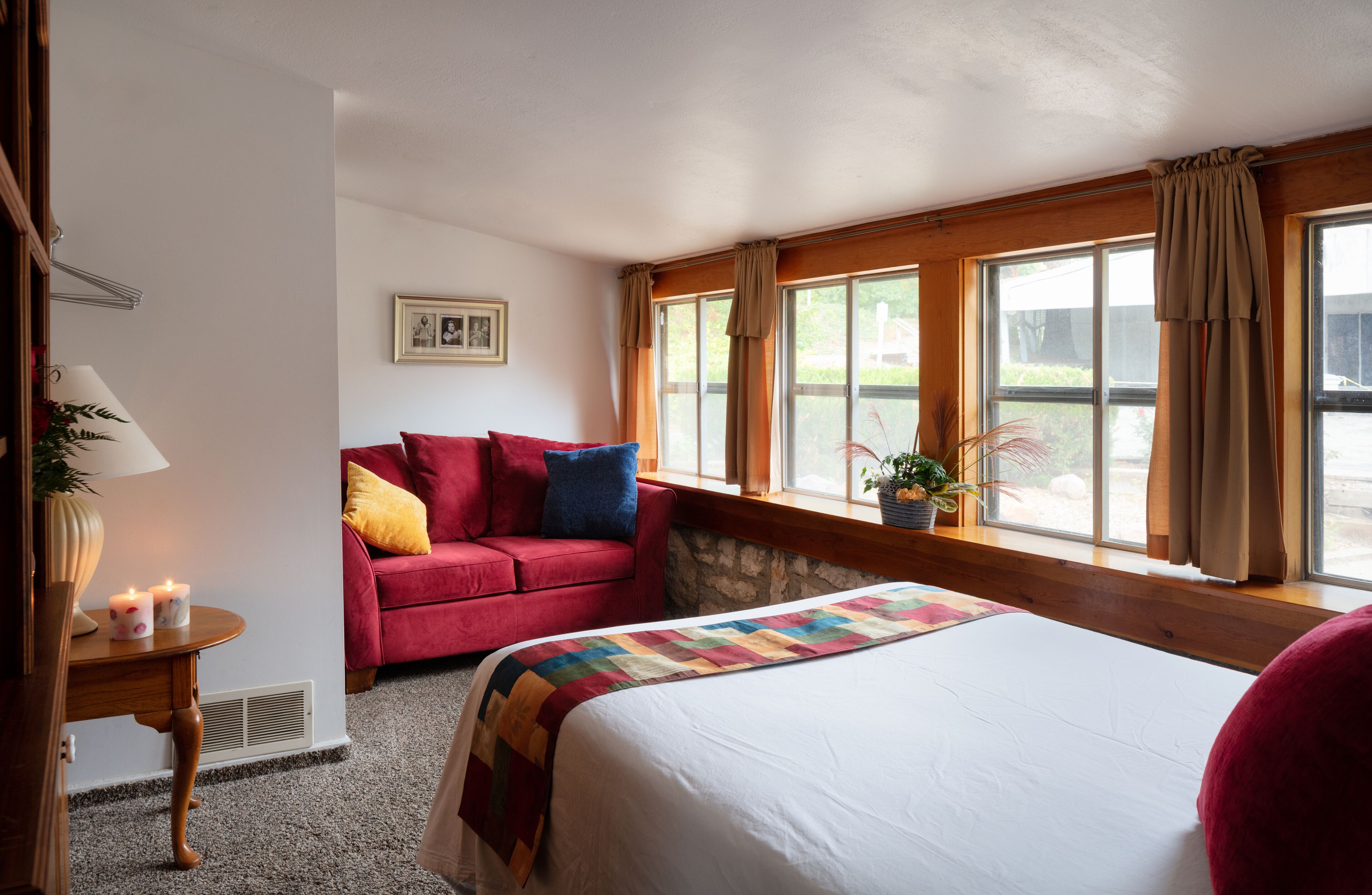 Ein Hotelzimmer mit einem Bett, einem roten Sofa, einem Fenster mit Vorhängen, einem kleinen Tisch mit einer Lampe und einem gerahmten Bild an der Wand.