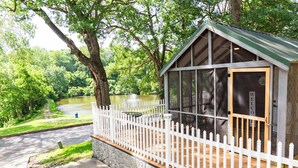 A wooden cabin with a white picket fence, a stone foundation, and a view of a lake.
