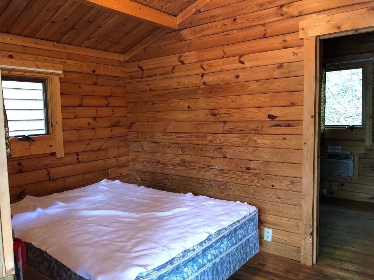 A wooden cabin room with a bed, a window, and a door leading to another room.