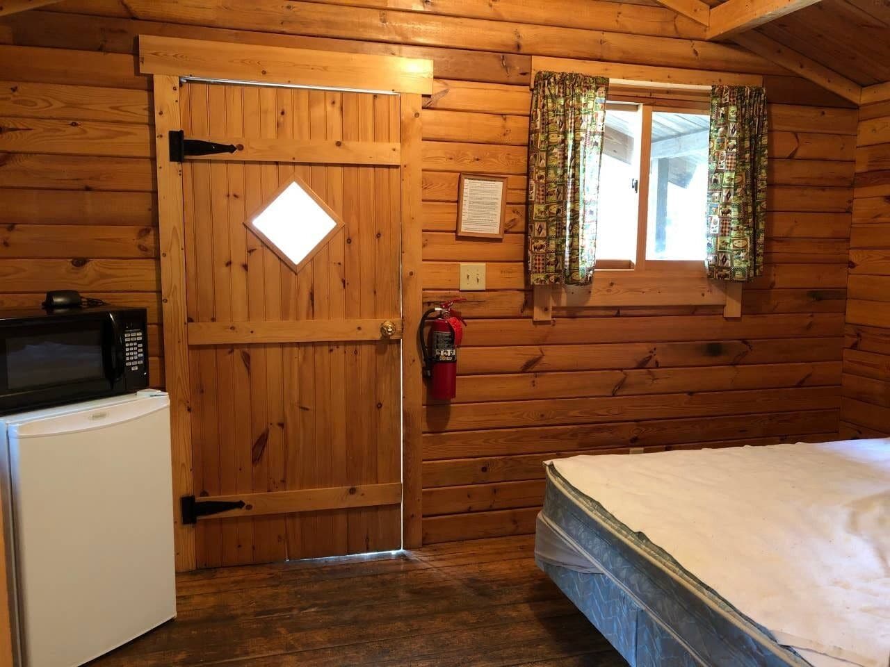 Camera di una baita in legno con un letto, un forno a microonde, un frigorifero, una porta e una finestra con tende.
