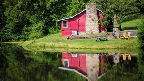 A red cabin with a stone chimney, a picnic table, and two wooden chairs by a calm lake, surrounded by lush greenery.