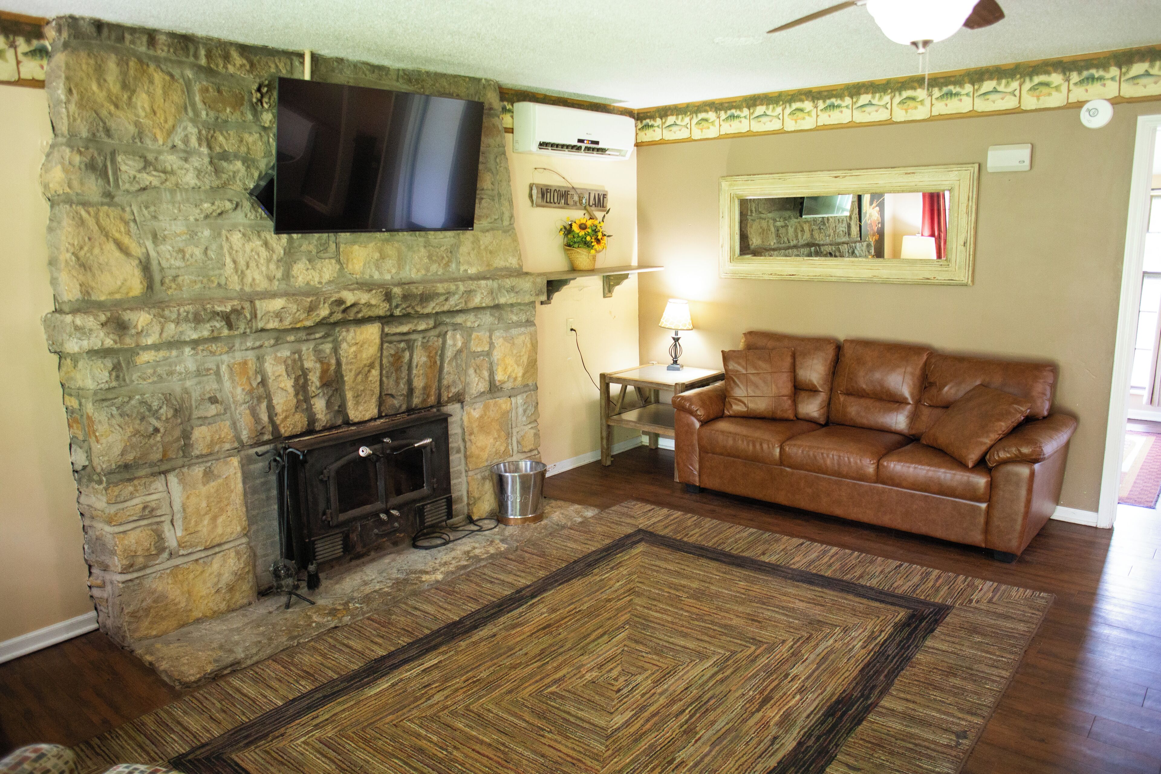 A living room with a stone fireplace, a leather sofa, a flat-screen TV mounted on the wall, and a patterned rug.