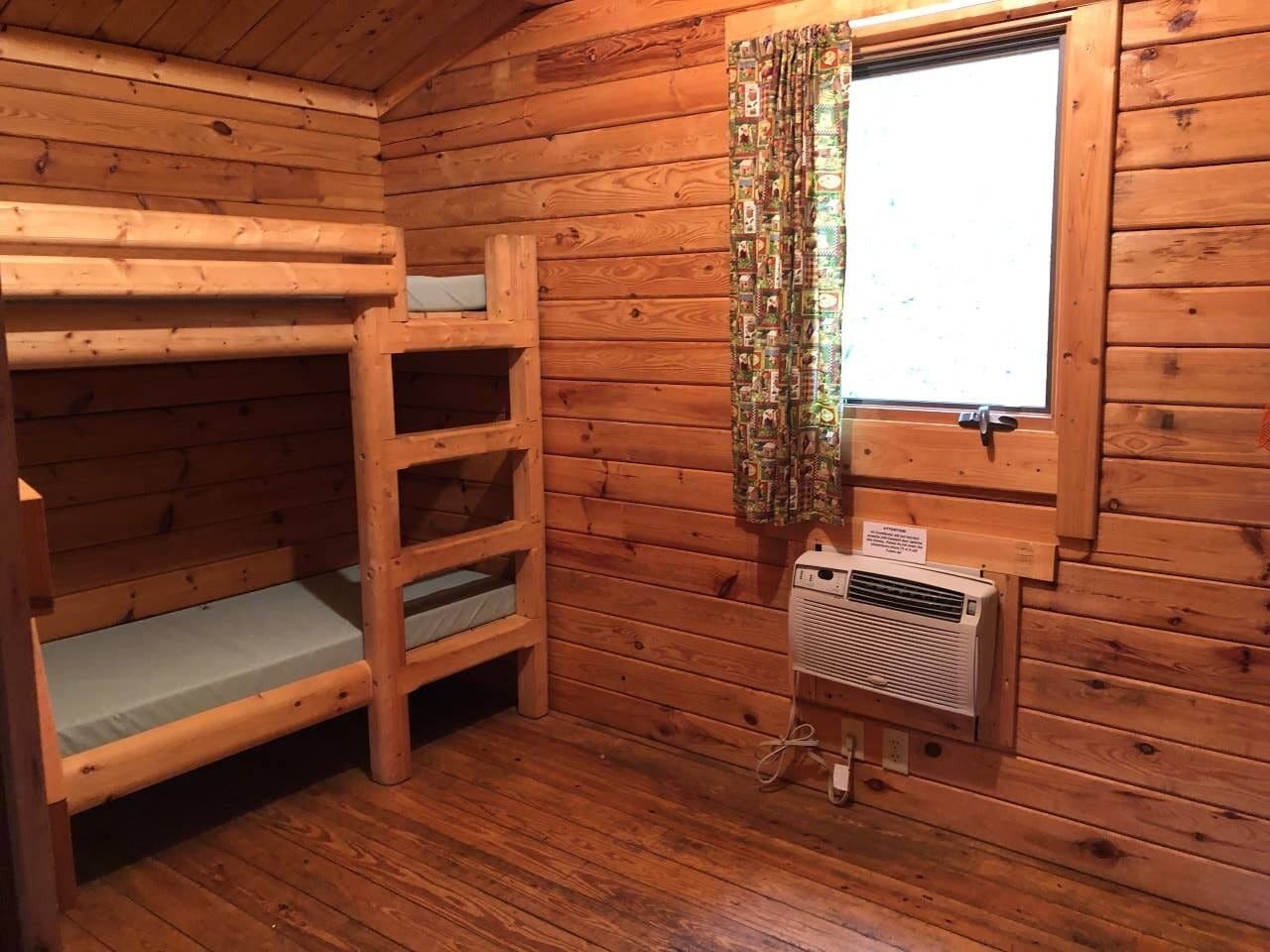 A wooden cabin room with bunk beds, a window with curtains, and a wall-mounted heater.