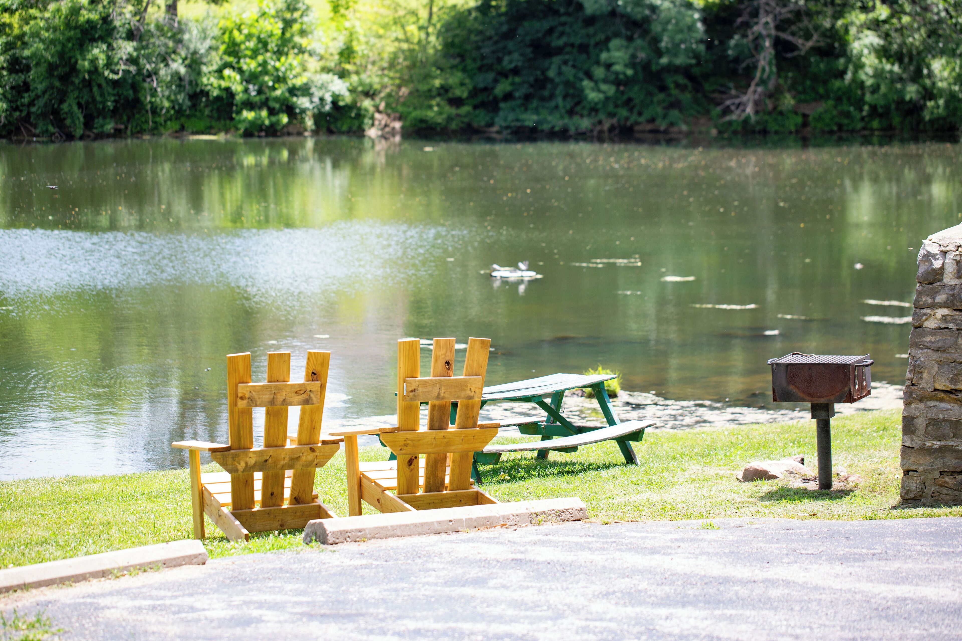 A lakeside setting with wooden chairs and a picnic table, a stone post with a mailbox, and a calm water body.