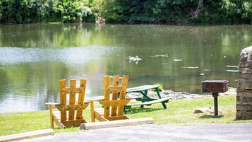 A lakeside setting with wooden chairs and a picnic table, a stone post with a mailbox, and a calm water body.