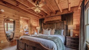 Un dormitorio de cabaña de madera con una cama grande, un ventilador de techo, un espejo y una mesita de noche con una lámpara.