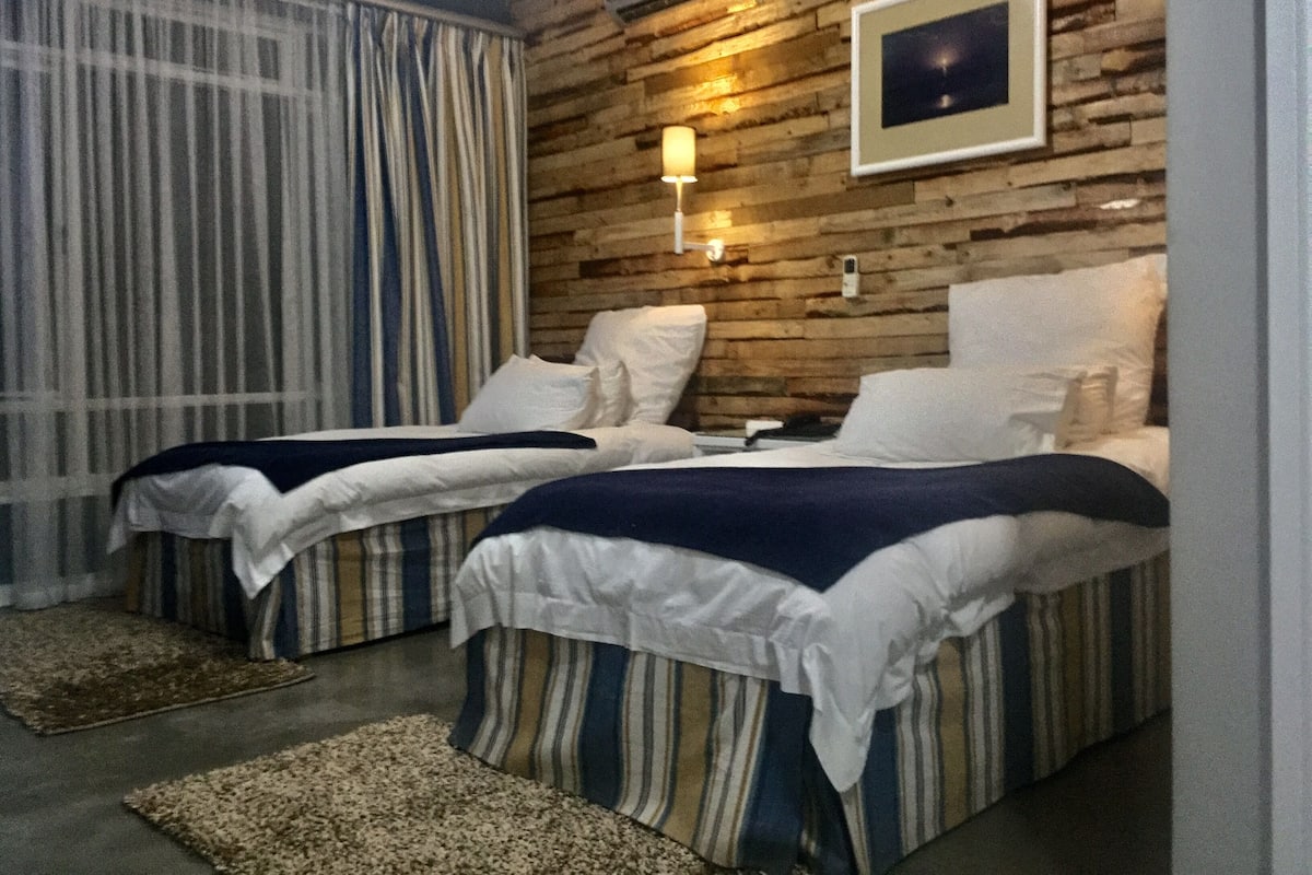 A hotel room with two beds, striped bedding, a wooden wall, a framed picture, and a window with curtains.