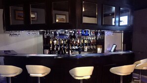 Zona de bar con mostrador negro, cuatro taburetes blancos y una variedad de botellas y vasos en estanterías.