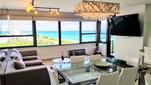 Una sala de estar moderna con una mesa de centro de vidrio, un sofá y sillones. Hay un televisor grande en la pared y una vista al océano a través de amplias ventanas.