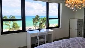 Un dormitorio con escritorio, cama y vistas a la playa y al océano a través de amplios ventanales.