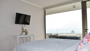 A bedroom with a bed, a mounted TV, a small table with a decorative horse statue, and a large window offering a city view. - Kennedy Dream - Indoor Pool, AC, next to Shopping (Vitacura)