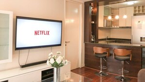 A modern living room with a flat-screen TV displaying 'NETFLIX', a glass coffee table, a vase with flowers, and a kitchen area with a bar stool. - Kennedy Dream - Indoor Pool, AC, next to Shopping (Vitacura)