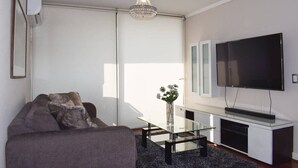 A modern living room with a grey sofa, a glass coffee table, a flat-screen TV, and a chandelier. - Kennedy Dream - Indoor Pool, AC, next to Shopping (Vitacura)