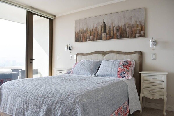 A bedroom with a large bed, a cityscape painting, a sliding glass door, and a white nightstand with drawers. - Kennedy Dream - Indoor Pool, AC, next to Shopping (Vitacura)