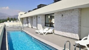 Une piscine sur le toit, agrémentée de chaises longues blanches, d’un mur en pierre et offrant une vue dégagée sur les montagnes en arrière-plan.