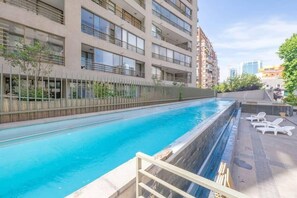 En swimmingpool som ligger intill en modern bostadsbyggnad med balkonger, omgiven av grönska och en klar himmel.