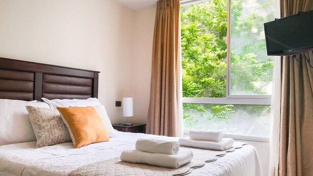 A neatly made bed with a quilt, pillows, and folded towels, a bedside table with a lamp, and a window with curtains. - Bellavista Spotlight 1BR Apt - w/Parking & A/C (Providencia)