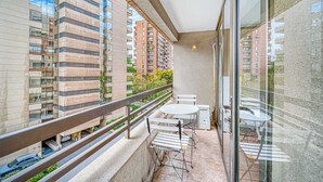 A balcony with a glass railing, a table, and chairs, overlooking a cityscape with modern buildings.