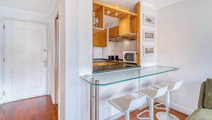 A compact kitchen with a glass bar counter, white bar stools, wooden cabinets, and a microwave.