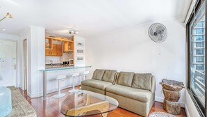 A modern living room with a glass coffee table, a beige sofa, and a kitchen area with white cabinets and a glass countertop.