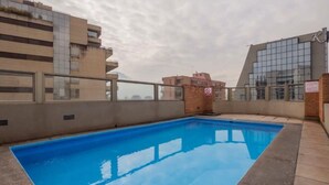 A rooftop swimming pool with a clear blue water surface, surrounded by a low brick wall and a metal railing.