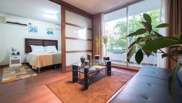 A hotel room with a bed, a glass coffee table, a sofa, a large window with curtains, and a plant. - Bellavista Serenity 1BR - w/Parking, AC & Terrace (Providencia)