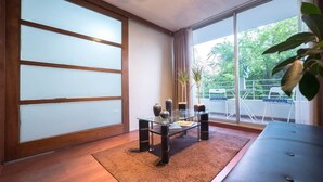 A modern living room with a glass-top coffee table, a dark sofa, and a view of a balcony with outdoor furniture. - Bellavista Serenity 1BR - w/Parking, AC & Terrace (Providencia)
