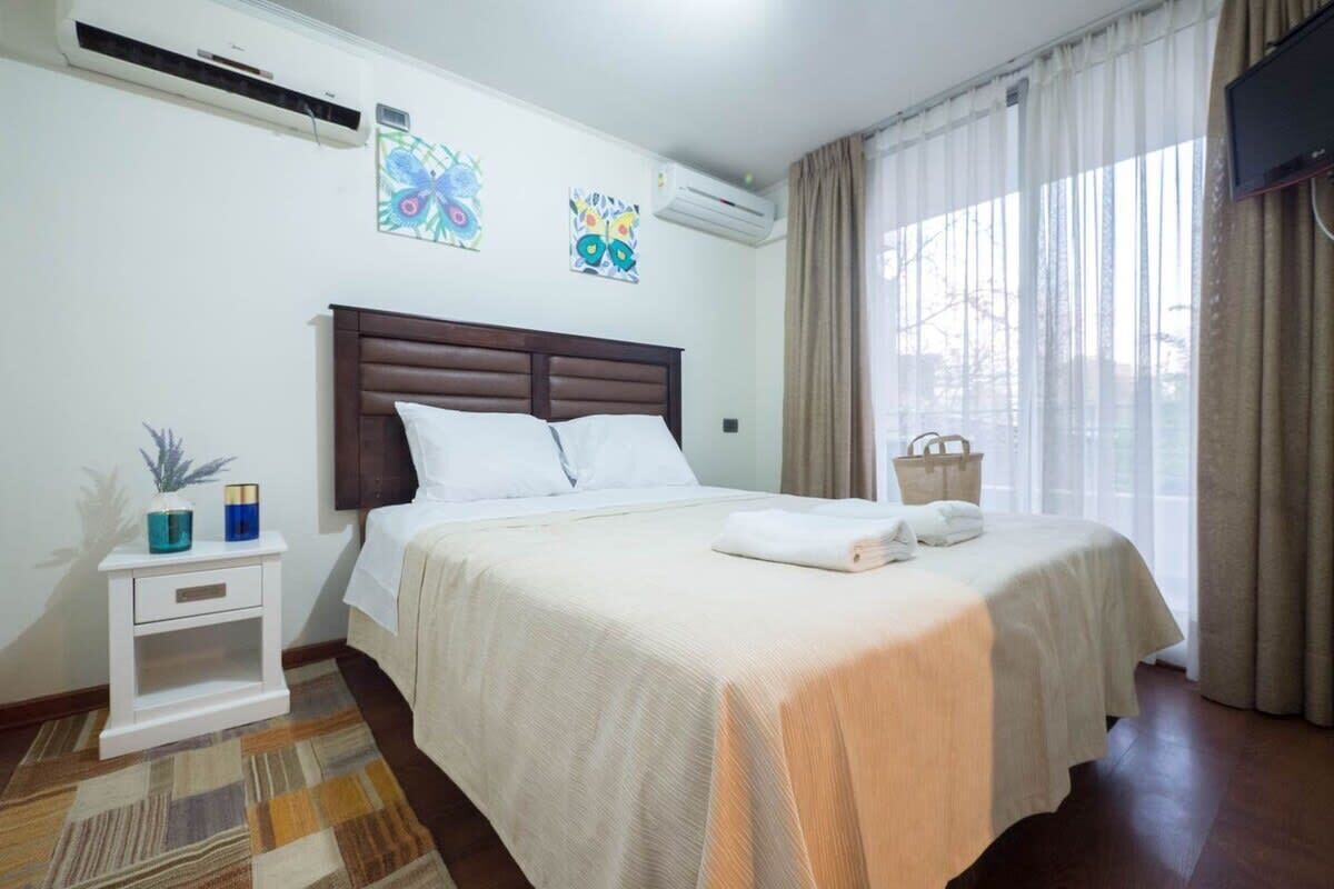 A hotel room with a large bed, a nightstand, a wall-mounted TV, and a window with curtains.