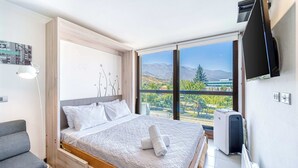 A bedroom with a bed, a gray chair, a TV, and a large window with a mountain view.