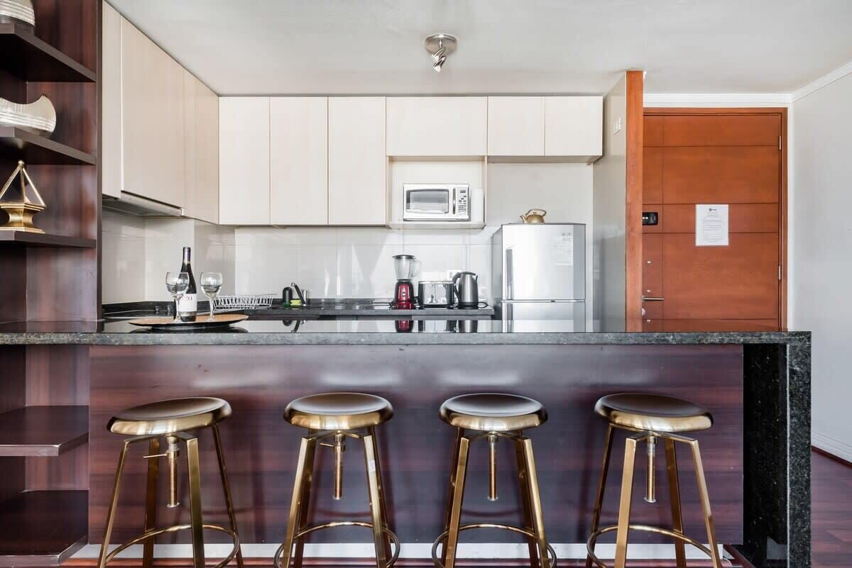 Een moderne keuken met witte kastjes, een magnetron, een koffiezetapparaat en een koelkast. Er staan drie barkrukken bij de bar.