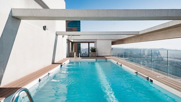 A rooftop swimming pool with clear blue water, surrounded by a modern architectural design and offering a view of the cityscape.