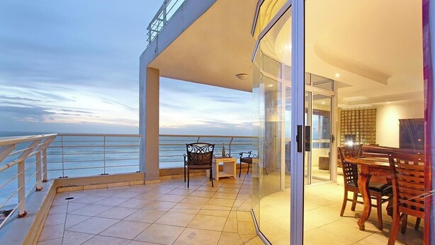 A balcony with a view of the sea, a dining area with wooden furniture, and a glass door leading to an interior space. - Ocean View C602 bt HostAgents (Cape Town)